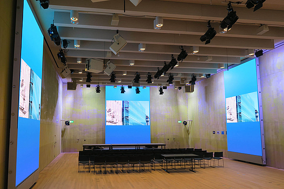 Großer Veranstaltungssaal mit Sitzplätzen und drei schmalen Video Walls