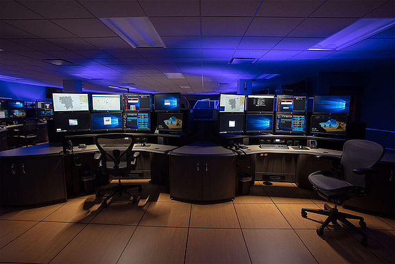 Blau beleuchteter Control Room mit mehreren Arbeitsplätzen und zahlreichen Bildschirmen